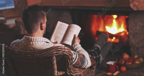 Man Reading a Book by the Fireplace. SLOW MOTION 4K. Young man reading a book by the warm fireplace decorated for Christmas. Relaxed holiday evening concept.