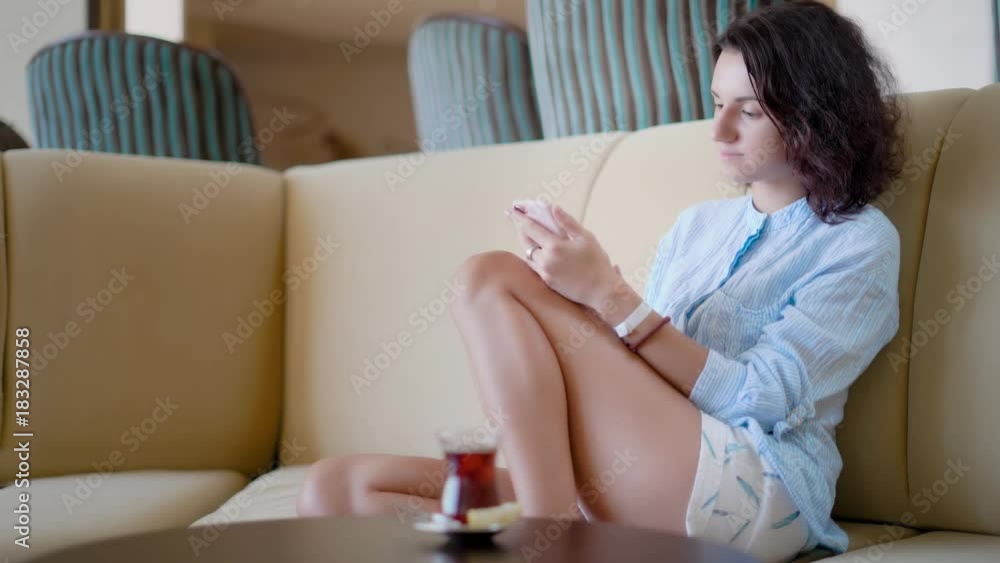 young pretty woman with short wavy hair is sitting on a couch and looking pictures in a smartphone young pretty woman with short wavy hair is sitting on a couch and looking pictures in a smartphone