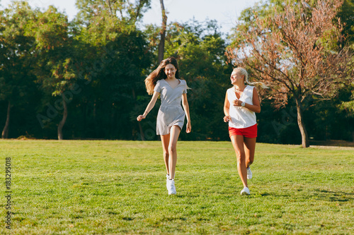 Happy young daughter with long brown hair and her mother running together at the park, sport in summer sunny time. Woman and girl outdoors. Family, lifestile concept.