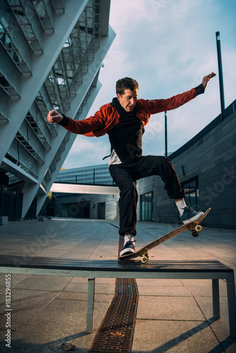 young skateboarder in stylish outfit balancing with board on bench