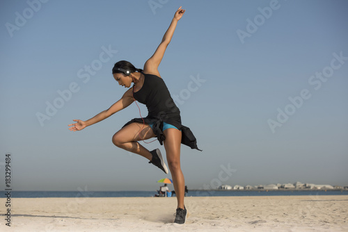 Beautiful Biracial woman by the ocean on a clear Summer morning does an expressive dance move wearing shorts and sports top as she listens to music on her headphones 