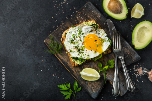 toast with avocado, spinach and fried egg on wooden cutting board, black background