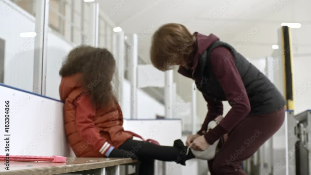 Zoom in of female coach tying figure skates for little girl and discussing something with her before training on indoor ice rink Zoom in of female coach tying figure skates for little girl and discussing something with her before training on indoor ice rink