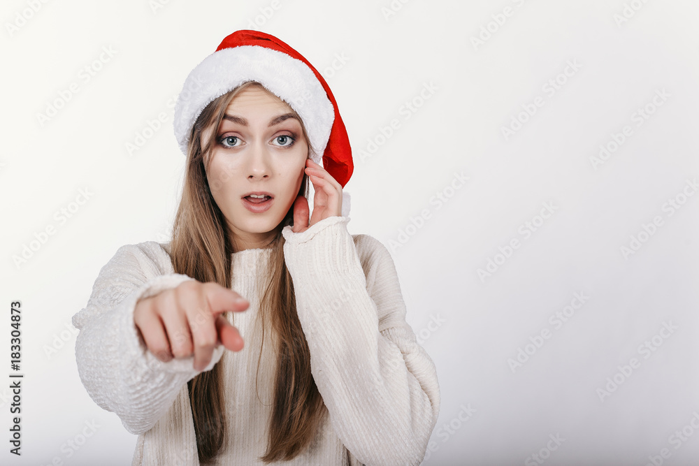 Woman in Santa hat pointing at camera by her finger. Girl suprised by somthing. Pretty young model with blonde long hair, wide eyebrows and blue eyes posing on camera in studio. isolated. Woman in Santa hat pointing at camera by her finger. Girl suprised by somthing. Pretty young model with blonde long hair, wide eyebrows and blue eyes posing on camera in studio. isolated.