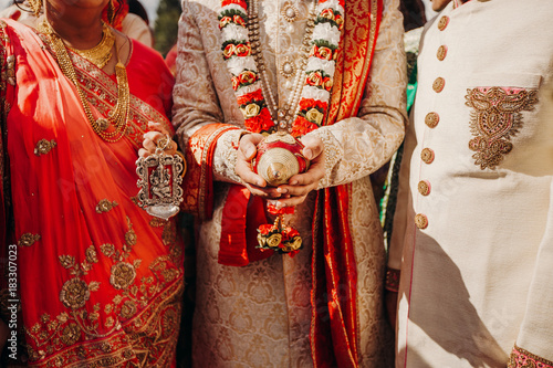 Hindu groom dressed in white sherwani embroidered with gold and perls holds a symbolic nut standing between the parents