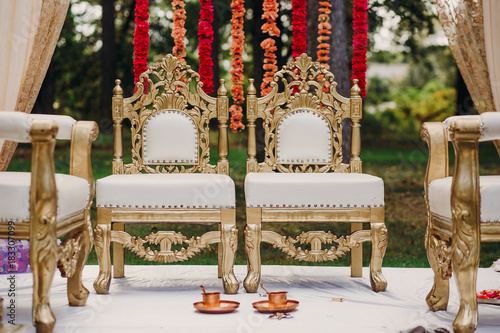 Bronze cups with spoons stand on the floor before golden chairs prepared for Hindu wedding photo