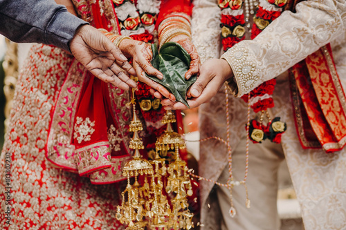 Indian bride and groom dressed in traditional shewrani, lehenga and with flower garlands on their necks hold green leaf during the Saptapadi wedding ceremony