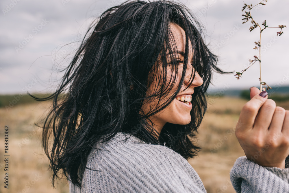 Fototapeta premium Side view portrait of happy brunette young woman wearing gray sweater being playful and carefree with beautiful smile and windy hair posing on landscape. Lifestyle concept. Cover idea mood.