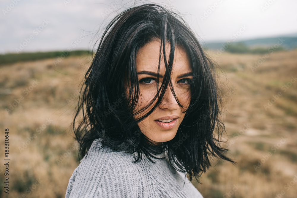 Fototapeta premium Horizontal portrait of romantic brunette woman posing against nature, meadow background with windy hair. People and lifestyle concept. Sensual natural female posing during travel. Cover idea mood.