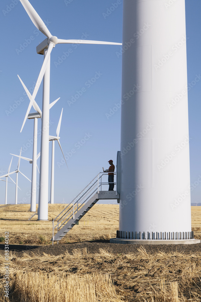 A wind farm technician standing and using a laptop at the base of a turbine on a wind farm in open countryside at Palouse. A wind farm technician standing and using a laptop at the base of a turbine on a wind farm in open countryside at Palouse.