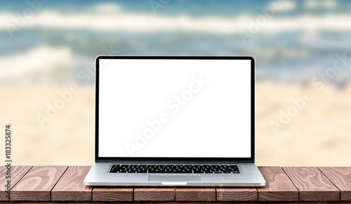 Modern laptop with empty white screen on wooden table against blurred beach bacmground photo