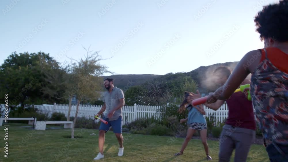 Woman with cylinder tube toy running towards her friends and spraying water. Group of friends playing with water gun toys and spraying each other outside in the backyard garden. Woman with cylinder tube toy running towards her friends and spraying water. Group of friends playing with water gun toys and spraying each other outside in the backyard garden.
