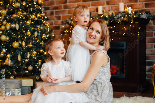 Cheerful happy woman with two daughters sitting on the floor in the living room near christmas tree. New Year's interior. Pretty mother with two little girls in dresses