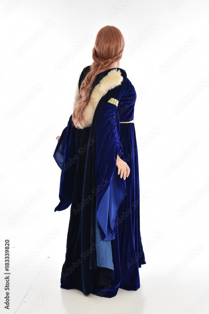 full length portrait of girl wearing long blue velvet gown and fur lined cloak, standing pose facing away from camera on white background.