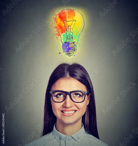 Portrait of a woman with glasses and creative idea looking up at colorful light bulb on gray background. Inspiration concept 