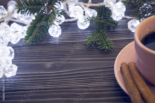 A dark wooden Christmas background with white fairy lights and fir tree branches at the top. A cup of coffee with cinnamon sticks on a saucer on a table. Close up. Copyspace