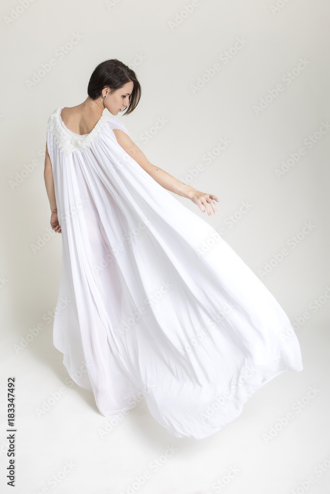 Slim fit caucasian brunette woman in white long dress in studio. Weightless fabric flowing, flying around girl in motion. Sensual soft portrait. White neutral background Slim fit caucasian brunette woman in white long dress in studio. Weightless fabric flowing, flying around girl in motion. Sensual soft portrait. White neutral background
