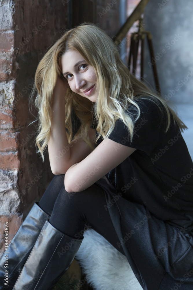 Fototapeta premium Cute blonde caucasian woman sitting in loft grunge trendy minimalistic interior, smiling cheerfully