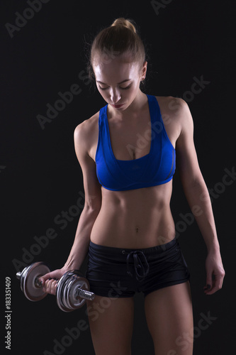 Beautiful brunette caucasian young woman with fit athletic body in sportswear do sport exercise with heavy dumbbell pumping up muscles on her hands. Neutral dark background