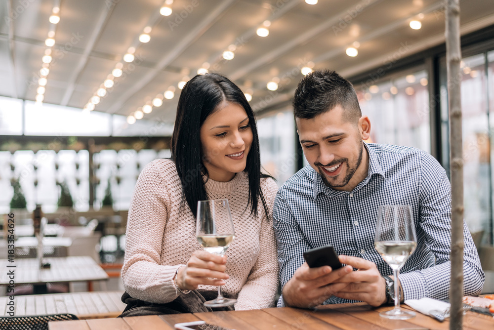 Portrait of happy young man and woman sitting together at a restaurant and looking at a mobile phone.