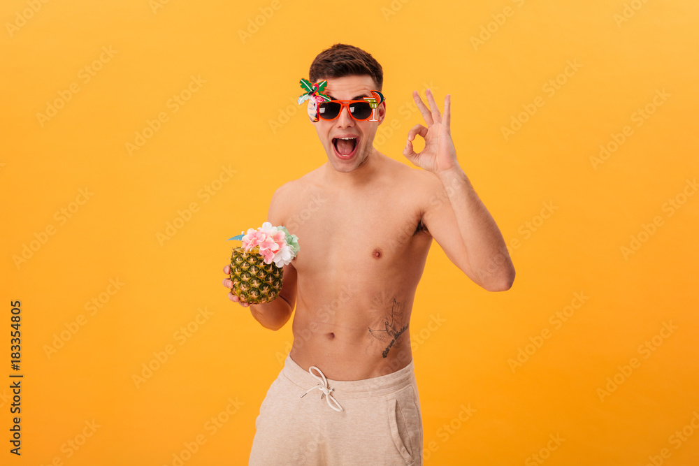 Fototapeta premium Happy naked man in shorts and unusual sunglasses holding cocktail