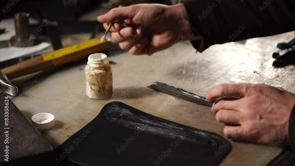 Leathersmith is applying glue on a leather parts of the zipper with a special brush. He works at the table in the workshop. Leathersmith is applying glue on a leather parts of the zipper with a special brush. He works at the table in the workshop.