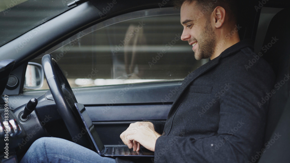 Attractive smiling man typing laptop computer while sitting inside his car outdoors Attractive smiling man typing laptop computer while sitting inside his car outdoors