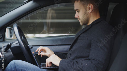 Attractive young man using tablet laptop computer while sitting inside his car outdoors