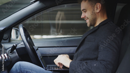 Attractive smiling man typing laptop computer while sitting inside his car outdoors © silverkblack