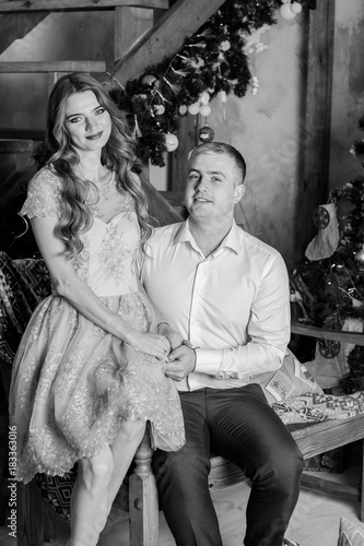 New year holiday portrait of a man and woman surprising him with a Christmas gift. Couple celebrating Christmas together. Beautiful young couple is celebrating at cozy romantic place.