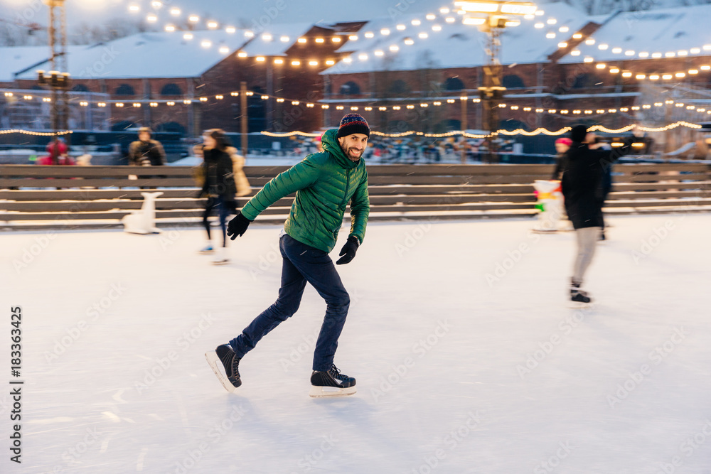 Joyful bearded man practices going skating on ice ring, has cheerful expression, smiles happily, demontrates his professionalism. Active sporty male in green jacket go in for winter sport