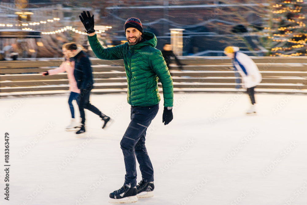 Obraz premium Happy male waves with hand as notices his friend on skating ring, have desire to skate together, rejoices meeting, has good time and mood, entertains himself. People and winter activities