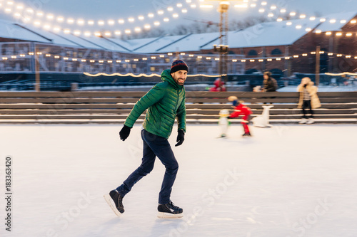 Professional male skater shows his skating talents, being sure on ice ring, looks delightfully in camera, demonstrates his nice skills. Smiling happy man on skating ring. Leisure and professionalism