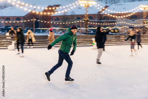 Outdoor shot of attractive man with beard, wears warm winter clothes, practices going skating on ice skate ring decorated with lights, has glad expression, enjoys his favourite action © VK Studio