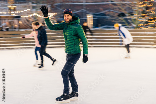 Happy male waves with hand as notices his friend on skating ring, have desire to skate together, rejoices meeting, has good time and mood, entertains himself. People and winter activities