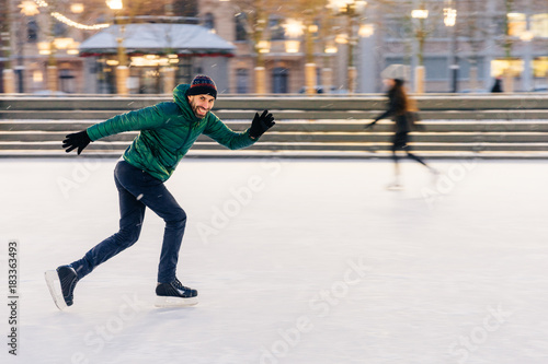Happy active sporty male involved in winter activities, demonstrates his skating skills on Christmas decorated ice ring, being in movement, has active lifestyle, enjoys his favourite hobby
