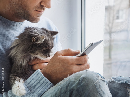 Young man sits on the windowsill, holds a beautiful, fluffy kitten on his lap and reads news on his mobile phone