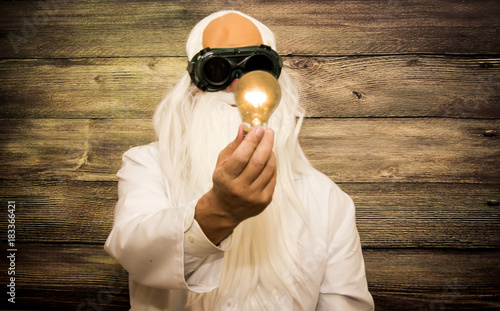 Crazy scientist elderly man holding an inexplicably lit bulb with some strange glasses on wooden background photo