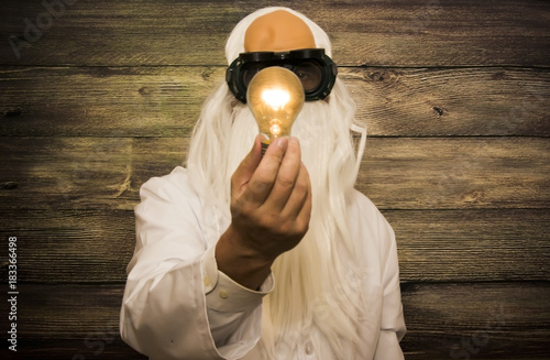 Crazy scientist elderly man holding an inexplicably lit bulb with some strange glasses on wooden background photo