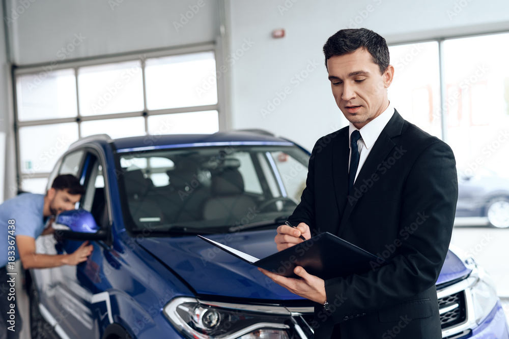 A car salesman is posing for a camera near buyers who are choosing a new car. A car salesman is posing for a camera near buyers who are choosing a new car.