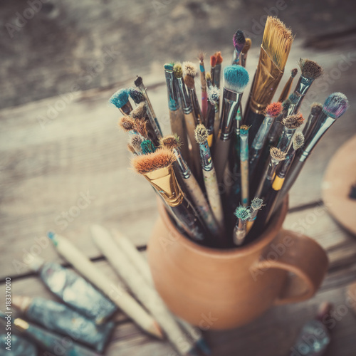 Paintbrushes in a jug from potters clay, wooden palette and paint tubes.