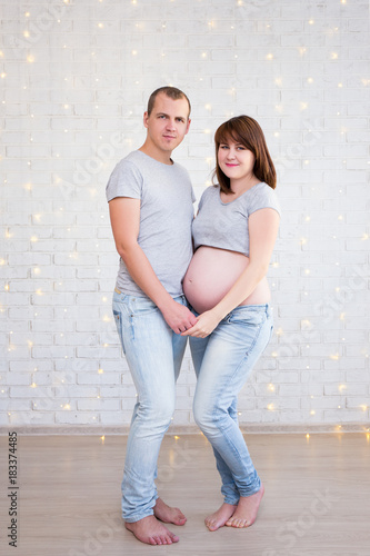 full length portrait of cute pregnant couple posing over white brick wall