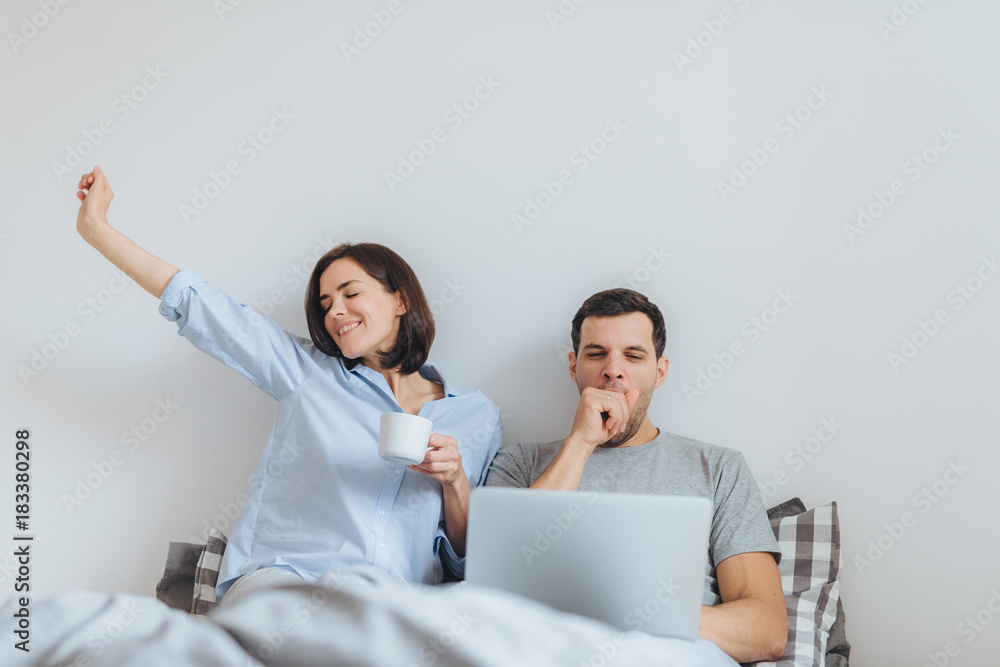 Glad female stretches in bed, drinks hot coffee and her husband works all night on laptop computer, yawns as wants to sleep, spend time together in bedroom, isolated over white background Glad female stretches in bed, drinks hot coffee and her husband works all night on laptop computer, yawns as wants to sleep, spend time together in bedroom, isolated over white background