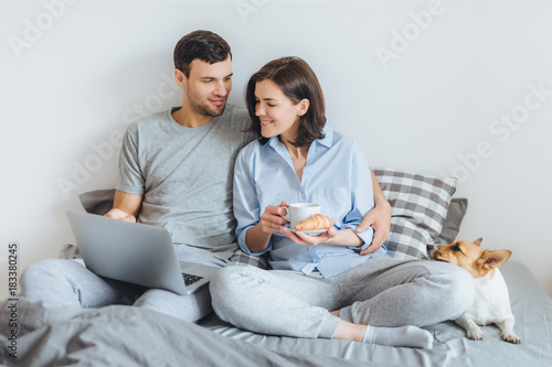 Affectionate couple in love embrace in bedroom, warch film online on laptop computer, have breakfast, enjoy domestic atmopshere and their favourite pet lies in bed. People, relatioships concept