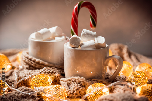 Christmas background with cocoa or coffee with marshmallows and candy in a white cup on a brown knitted winter scarf and a glowing golden garland. Beautiful concept of home comfort and warmth