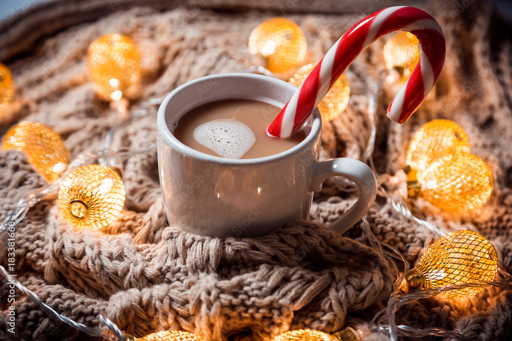 Christmas background coffee in a white cup with candy on a brown knitted winter scarf and a glowing golden garland. Beautiful home Holiday drink Christmas background coffee in a white cup with candy on a brown knitted winter scarf and a glowing golden garland. Beautiful home Holiday drink