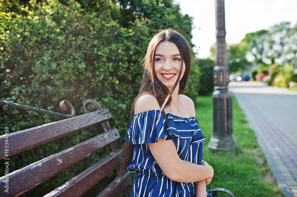 Gorgeous brunette girl sitting on bench at street of city wear on blue striped dress. Gorgeous brunette girl sitting on bench at street of city wear on blue striped dress.