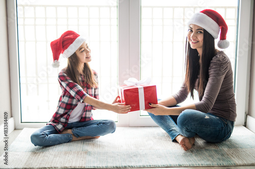 Happy mother and daughter wearing santa hats, sitting on floor and holding a gift box at home photo