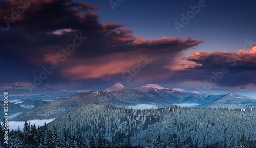 Panoramic view of dramatic sunset in the winter mountains. Wooded hills covered with snow, fog rising from valleys, colorful cloudy sky - this is impressive picture.