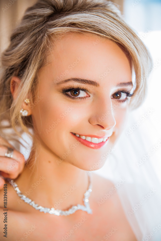 Portrait of the young beautiful smiling bride, her mother dressing up the necklace on neck of her daughter indoors